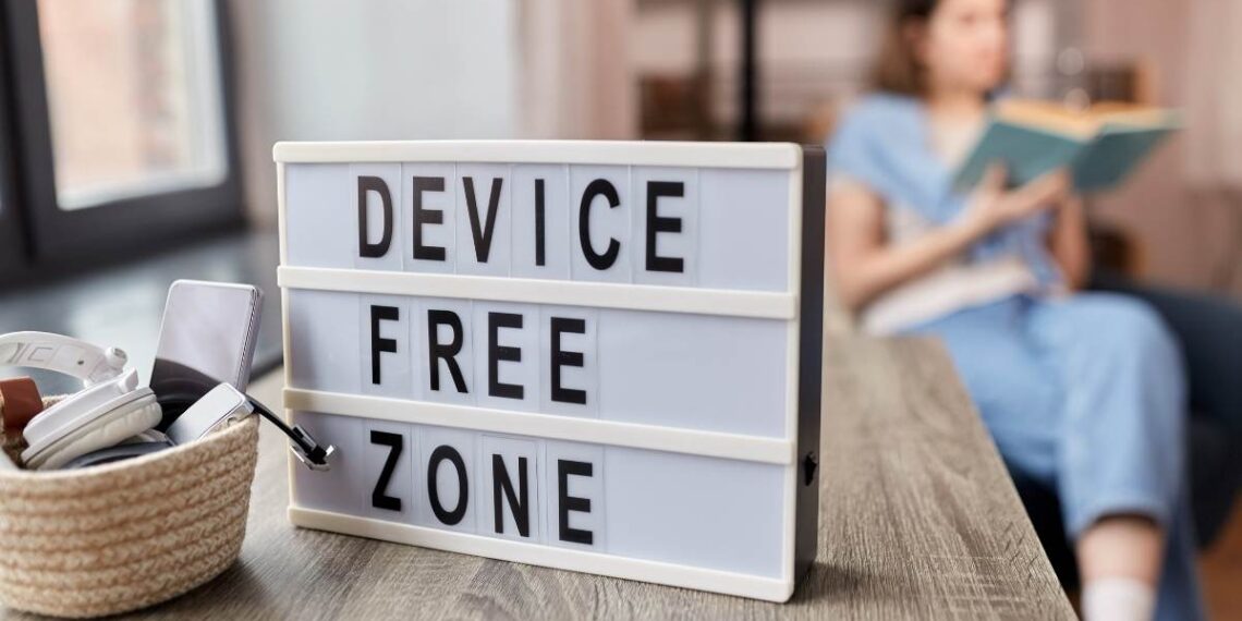 A device-free zone sign with a phone placed aside while a person reads a book in the background, illustrating simple way to spend less time on your phone and reduce screen time.