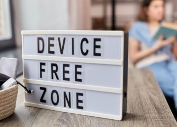 A device-free zone sign with a phone placed aside while a person reads a book in the background, illustrating simple way to spend less time on your phone and reduce screen time.