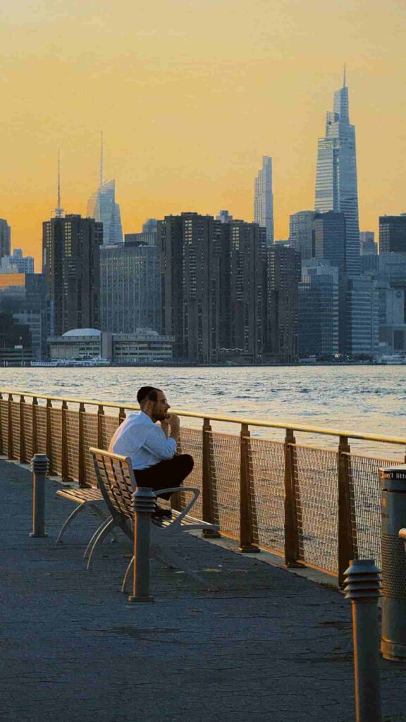 A person sitting alone by the waterfront, looking toward the city skyline at sunset