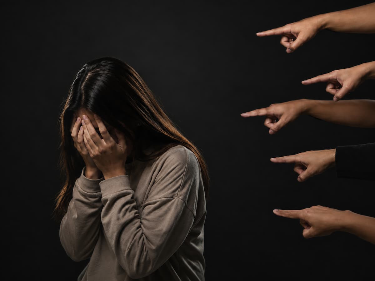 A woman hiding her face as several hands point at her, surrounded by criticism that makes her question her worth.